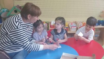 На изображении женщина в полосатой кофте общается с тремя детьми за круглым столом. Вокруг игрушки и учебные материалы.